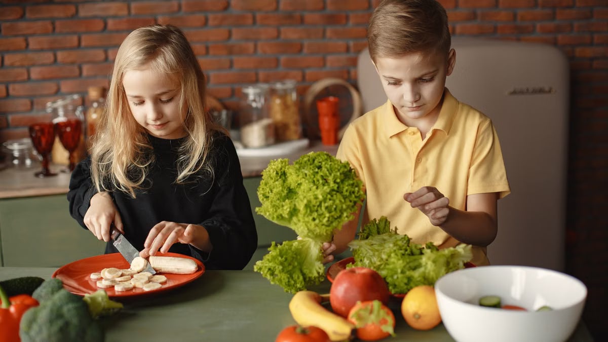 Focused little girl and boy sitting at desk with assorted vegetables and fruits while cooking in loft kitchen with brick wall